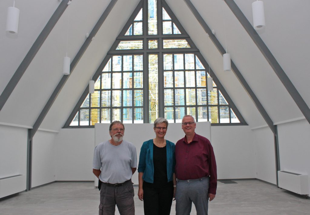 Hell und einladend: Presbyteriumsvorsitzender Jürgen Engelmann (v. l.), Pfarrerin Almuth Reihs-Vetter und Pfarrer Claus-Jürgen Reihs stehen in der umgebauten Immanuel-Kirche. Vor der leicht geschwungenen Rückwand unter dem Fenster des Glaskünstlers Günter Grohs werden zukünftig der Altar, das Taufbecken und der Ambo (Lesepult) ihren Platz haben. Foto: EKP/Oliver Claes