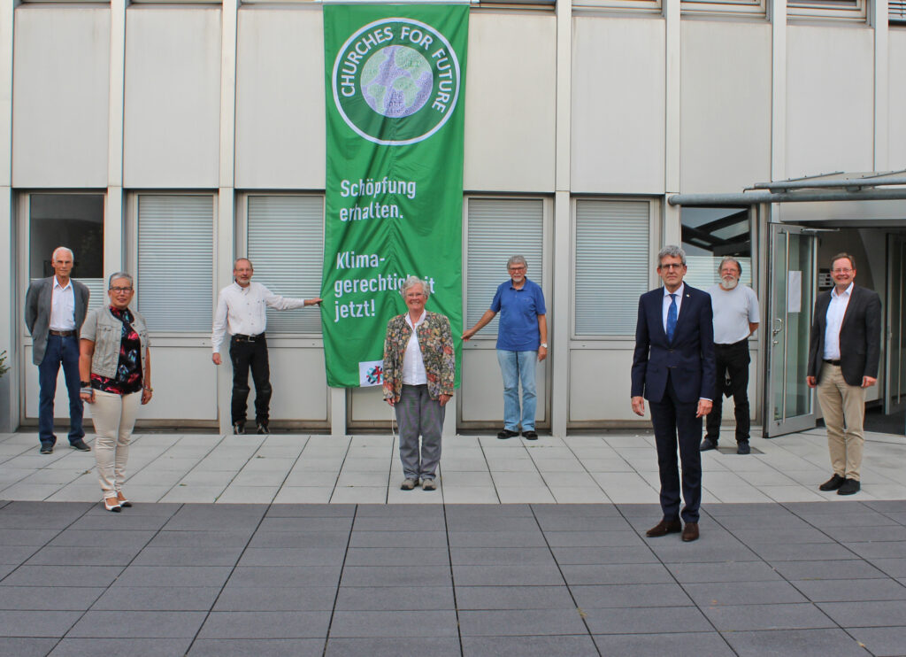 Rufen zum Einsatz für Klimagerechtigkeit auf: Die Mitglieder des Kreissynodalvorstandes des Evangelischen Kirchenkreises Paderborn (v. l.) Jürgen Scherhans, Evelyne Schubert, Wolfgang Neumann, Helga Weber-Kruck, Dr. Wilfried Hauenschild, Superintendent Volker Neuhoff, Jürgen Engelmann und Gunnar Wirth vor einer Fahne des Netzwerkes „Churches for Future“ am Haus der Evangelischen Kirche in Paderborn. Foto: EKP/Oliver Claes