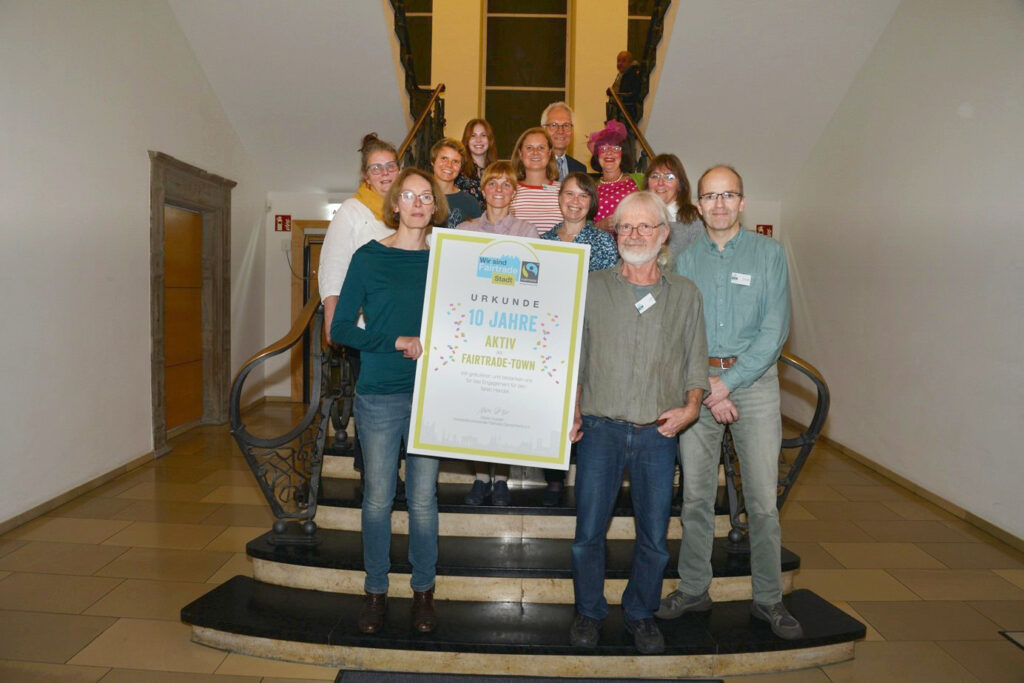 Die Initiative Faires Paderborn ist stolz auf die Auszeichnung „10 Jahre Fairtrade-Stadt Paderborn“ von Transfair Deutschland e. V.: Petra Holtgreve, Ludwig Feller und Ulrich Pietsch (erste Reihe: von links nach rechts), Heike Mahl, Christina Krengel, Martina Gamm, Tanja Steins (zweite Reihe: von links nach rechts), Lara Schröder, Jenny Fromme (dritte Reihe: von links nach rechts), Lara Brundiers, Stellvertretender Bürgermeister Martin Pantke und Moderatorin Else Mögesie (vierte Reihe: von links nach rechts). Aus der Steuerungsgruppe fehlen: Mechthild Goldstein, Stefan Pfeifer, Anke Reimer, Britta Bertelsmeier und Niklas Voß. Foto: Stadt Paderborn/Anna-Marina Vogt