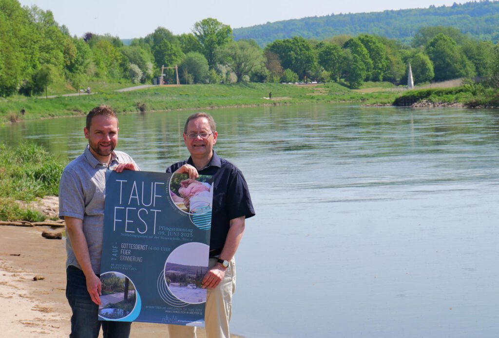 Die Pfarrer stehen mit dem Plakat zum Tauffest am Ufer der Weser in Höxter.