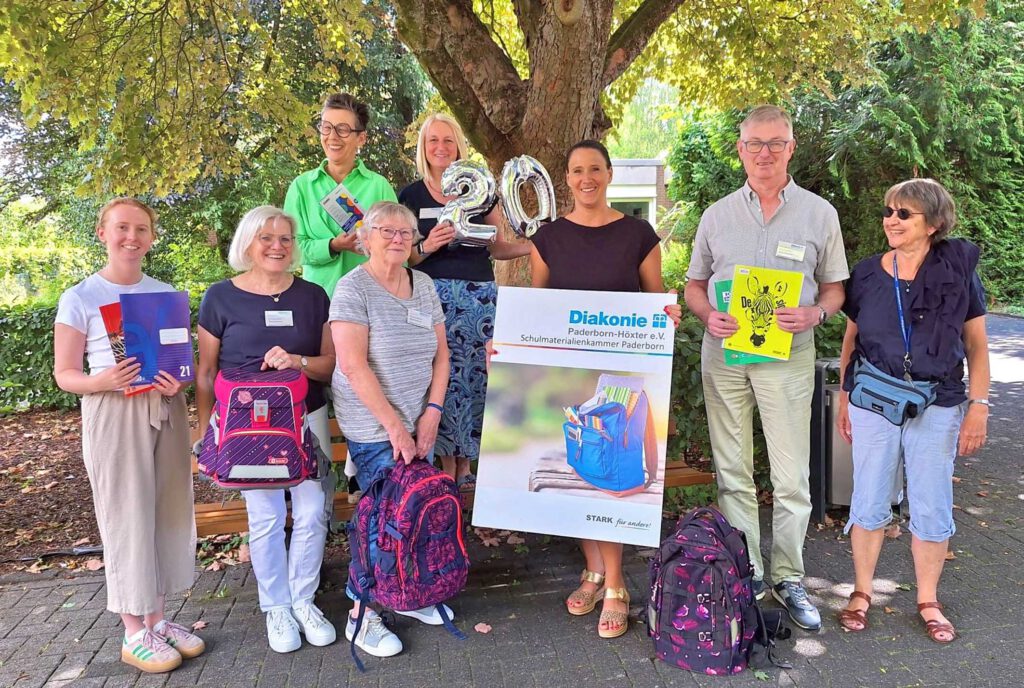 Mitarbeitende der Schulmaterialienkammer mit Schultaschen und einem Plakat.
