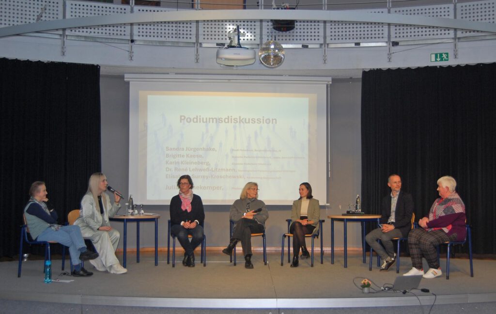 Die Teilnehmenden der Podiumsdiskussion sitzen auf der Bühne im Veranstaltungsraum.