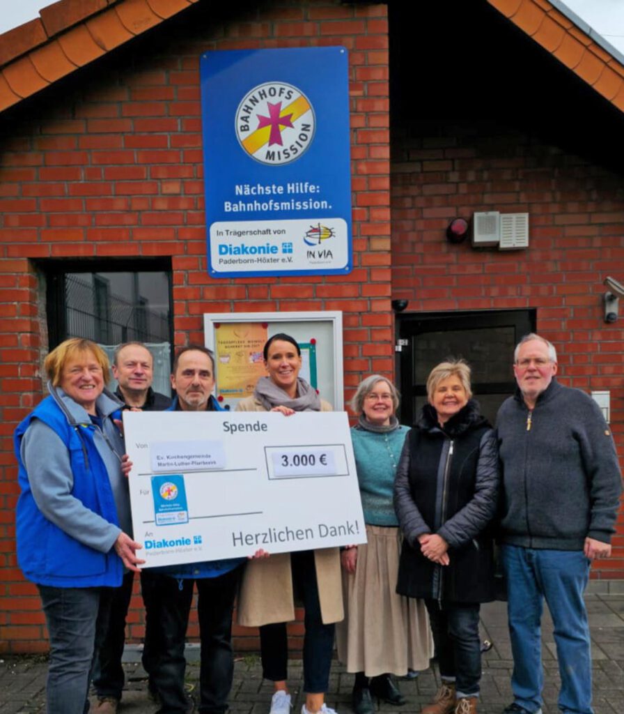 Mitarbeitende des Martin-Luther-Pfarrbezirks und der Bahnhofsmission stehen vor dem Gebäude der Bahnhofsmission und halten einen symbolischen Spendenscheck.
