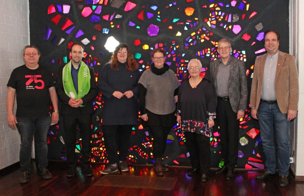 Sieben Personen stehen vor einem bunten Glasfenster in einer Kirche.