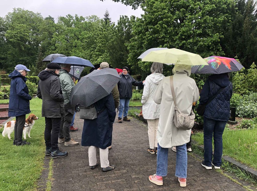 Menschen mit Regenschirmen hören einer Referentin beim Rundgang über einen Friedhof zu.