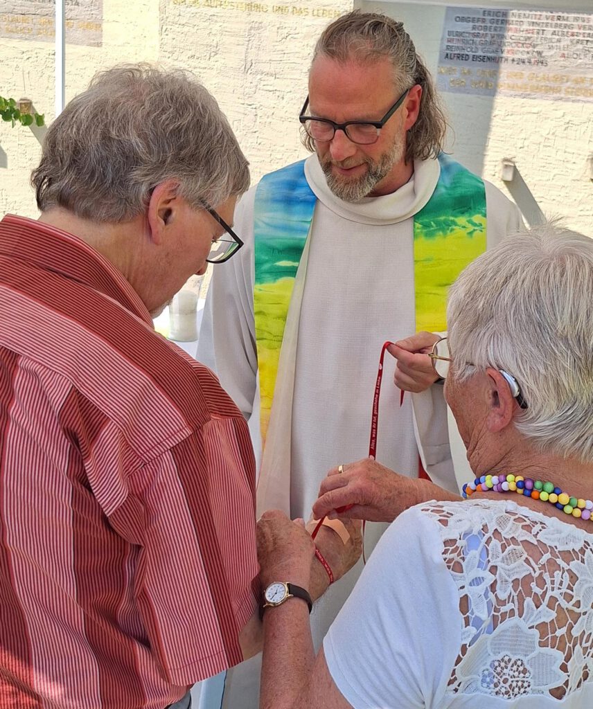 Ein Ehepaar wird von einem Pfarrer in einem Garten an einer Kirche gesegnet.