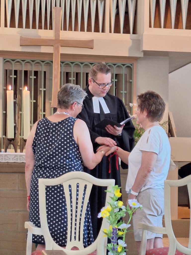 Zwei verheiratete Frauen werden von einem Vikar im Talar in einer Kirche gesegnet.