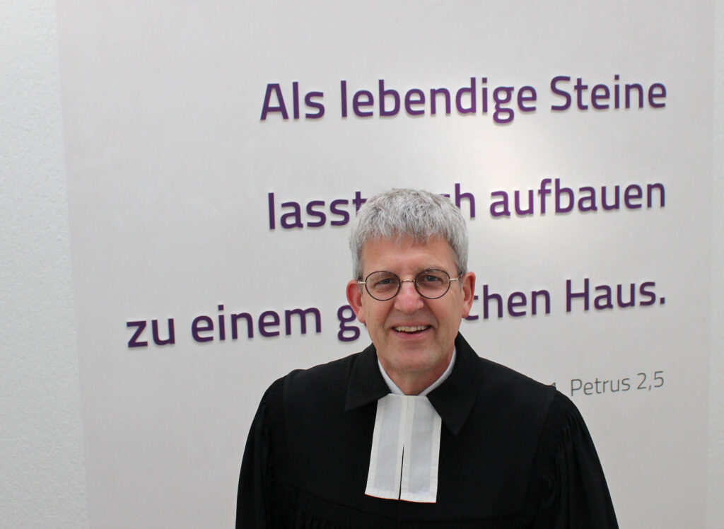 Der leitende Geistliche steht im Talar vor einer Wand, an der das biblische Leitwort des Kirchenkreises Paderborn angebracht ist.