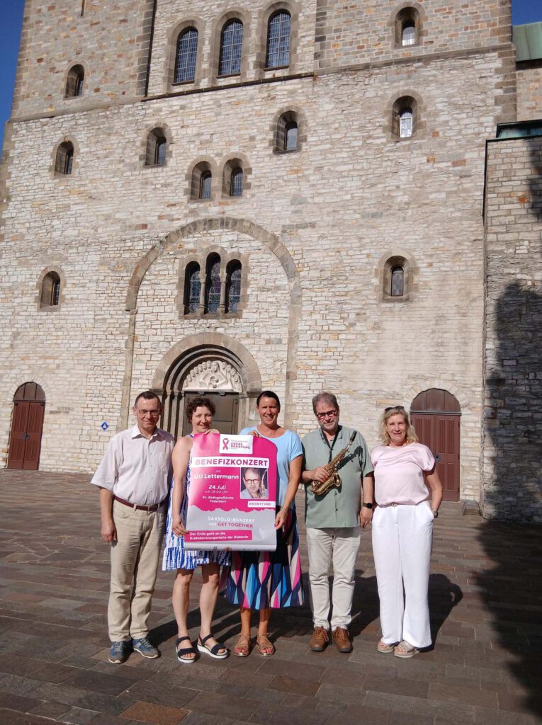 Menschen stehen vor einer Kirche und halten ein Plakat für ein Konzert in den Händen.