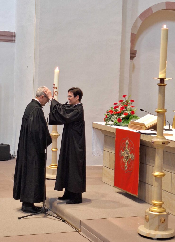 Eine Frau im Talar legt einem Mann im Talar in einer Kirche vor einem Altar segnend die Hände auf den Kopf.