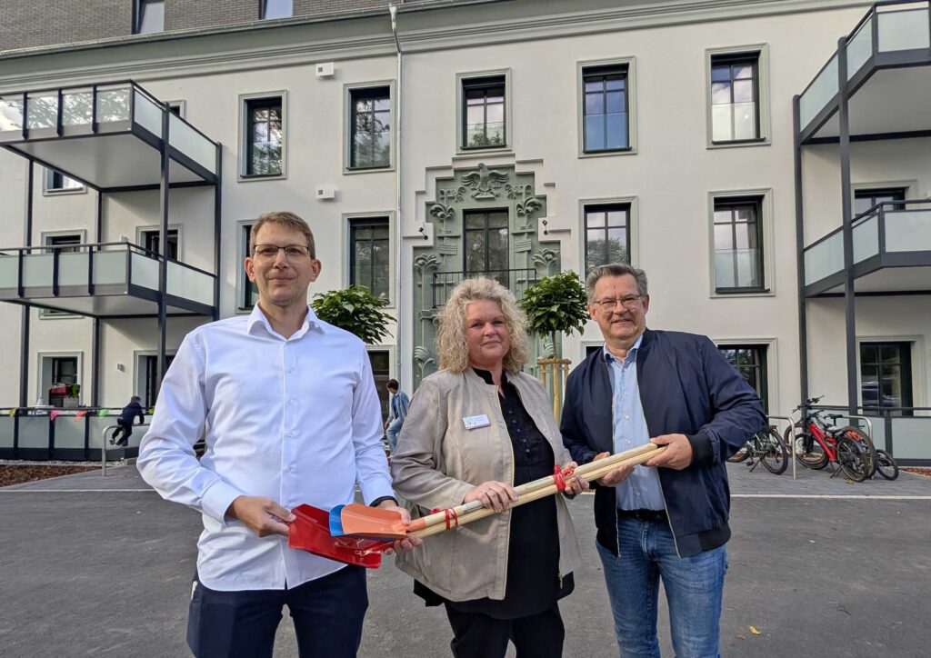 Drei Personen haben sich vor einem Kita-Gebäude aufgestellt und halten kleine Schaufeln in den Händen.